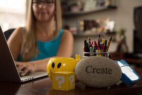 Cassidy sitting at a desk typing on a laptop with 'create' paperweight, a yellow box with question marks, and a yellow smiley face mug in foreground
